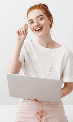 Horizontal shot of attractive redhead woman with bun hairstyle, touching rim of glasses and looking joyfully at camera while talking with coworker, working in brand new laptop, browsing in internet.